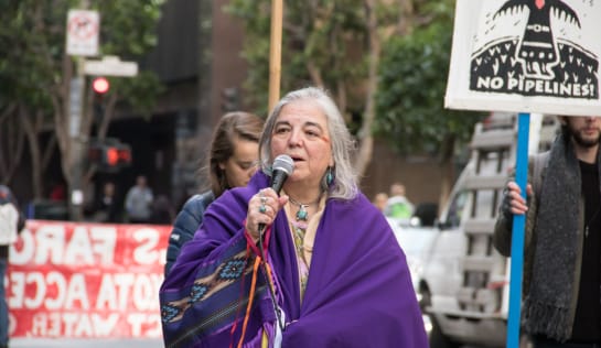 Eine ältere Frau mit einemlilafarbenen traditionellen Umhang spricht in ein Mikrofon anlässlich einer Demo gegen eine Ölpipline in ihrer Region in South Dakota
