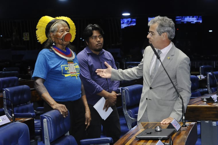 Raoni Metuktire mit gelbem Federkopfschmuck und T-Shirt und sein Enkel sprechen mit einem Senator im Senatssaal. Der Kayapó-Chief trägt die traditionelle runde Scheibe in der Unterlippe