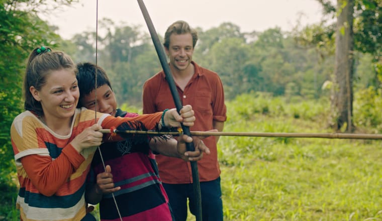 Checker Tobi schaut zu, wie seine Kollegin von einem Indigenen Jungen das Bogenschießen im Regenwald gezeigt wird