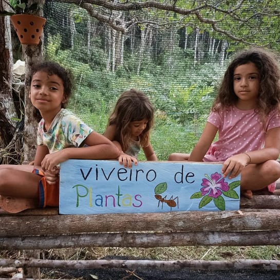 Drei Kinder halten ein blaues Plakat mit der Aufschrift Viveiro de Plantas (deutsch: Baumschule oder Gewächshaus). Sie haben einer Blattschneiderameise und eine violette Blume auf das Plakat gemalt