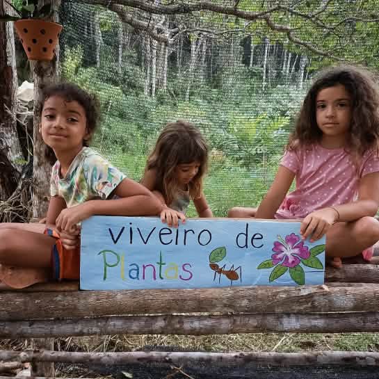Caoê, Lani und Nayani haben extra ein Schild gemalt Drei Kinder halten ein blaues Plakat mit der Aufschrift Viveiro de Plantas (deutsch: Baumschule oder Gewächshaus). Sie haben einer Blattschneiderameise und eine violette Blume auf das Plakat gemalt