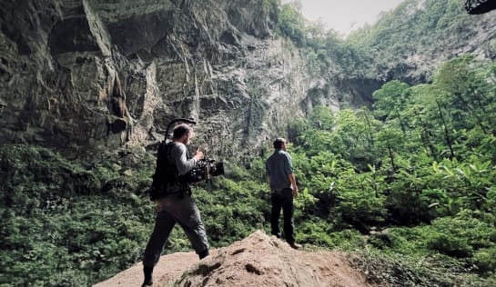 Tobi steht in einer mit Bäumen bewachsenen Höhle, umgeben von Felsen, hinter ihm ein Kameramann, der ihn filmt