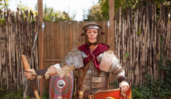 Tobi steht in einer Art Ritterrüstung mit Schild und Stock for einem Zaun