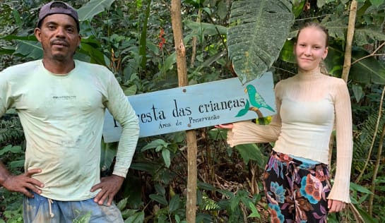 Rosenildo hat Annalena den „Wald der Kinder“ gezeigt. Dieses Waldstück wurde von den Kindern der Grundschule Steinkirchen und der Montessori Schule in Bamberg gespendet Ein Mann und eine junge Frau stehen im Wald, zwischen ihnen ein Schild mit der Aufschrift „Wald der Kinder“ (auf Portugiesisch)