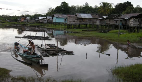 Fischer mit langen Holzstangen in einem Boot im See, am Ufer stehen Hütten auf Stelzen