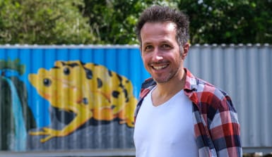 Willi Weitzel im Forschungslabor Panama Willi Weitzel steht vor einer Wellblechwand. Sie ist blau bemalt und zeigt den Goldenen Frosch von Panama. Es sind zwei gelbe Frösche mit schwarzen punkten, die aufeinander sitzen