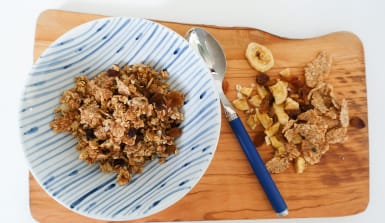 Granola Eine blau-weiße Schale gefüllt mit Müsli steht auf einem Olivenholzbrett, daneben liegen ein Esslöffel, Bananenchips und Flakes.