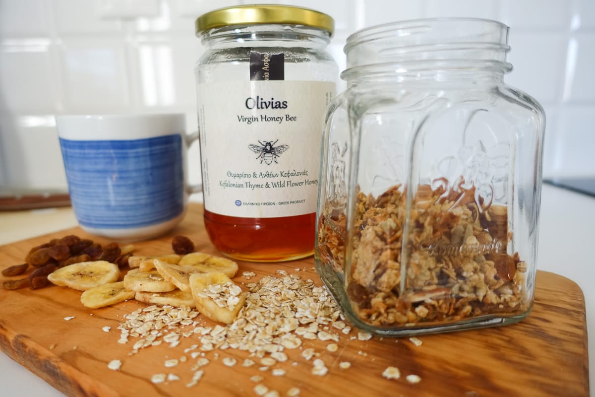Auf einem Olivenholtbrett stehen ein blau-weißer Becher, ein Honigglas und ein Schraubgas zur Hälfte gefüllt mit Granols. Auf dem Brett liegen Rosinen, Bananenchips und Haferflocken. 
