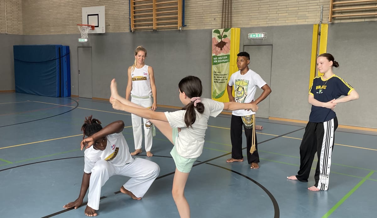 Figur aus der Kampfkunst: Caíque schützt sich vor dem Beinschwung der Schülerin 3 Personen schauen in einer Turnhalle eine Übung zwischen zwei Leuten zu, der Capoeira-Lehrer ist in der Hocke und chützt sich mit einem Arm vor dem ausgestreckten Bein einer Schülerin