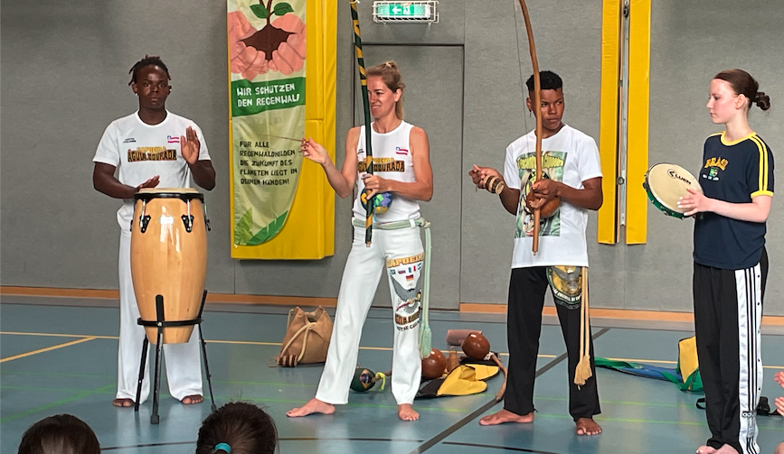 Wiedersehen in der alten Schule: Annalena (rechts) war natürlich dabei, als Daniel, Bianca und Caíque den Kindern des Gymnasiums Herzogenaurach einen Capoeira-Kurs geschenkt haben 2 junge Männer aus Brasilien und eine Frau in weißer Capoeira-Kleidung halten traditionelle Musikinstrumente hoch, die aus einem Ast, einer Saite und einer Kalebasse bestehen, einer der beiden Jungen schlägt eine Trommel, daneben eine junge Frau mit einem Tamburin. Im Vordergrund die zuschauenden Kinder in einem Halbkreis
