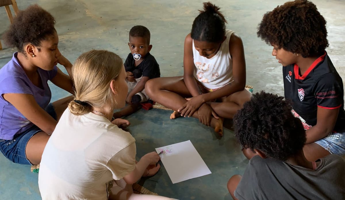 Unterricht mit Win-Win-Effekt: Die Kinder lernen Englisch, Annalena lernt Portugiesisch! Junge Frau sitzt mit 5 Kindern im Kreis auf dem Boden, in der Mittel liegt ein Blatt Papier