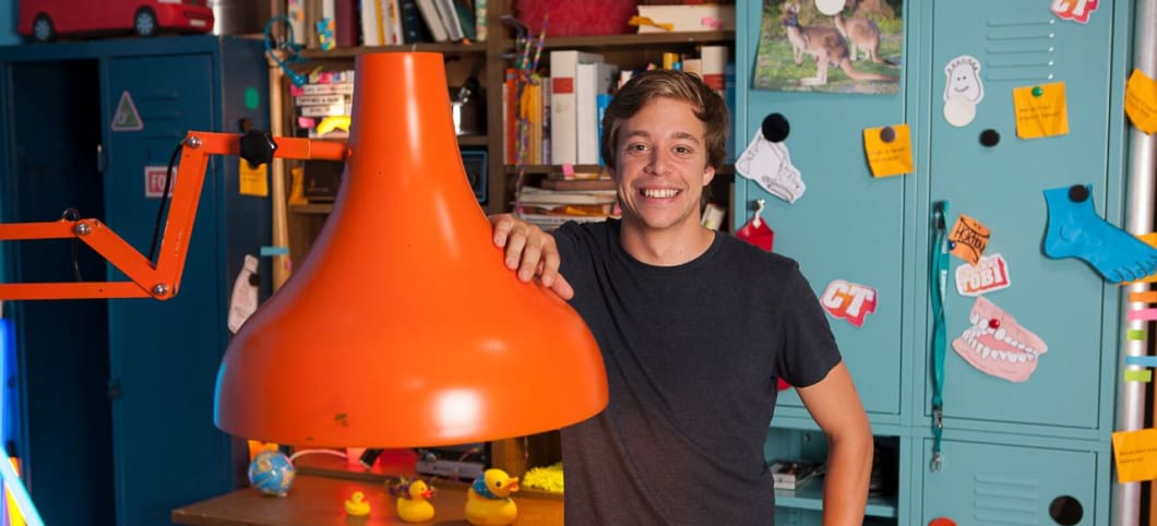 Tobi Krell in seiner TV-Checker-Bude. Von hier aus stürzt er sich in seine Abenteuer Tobias krell steht in einem Raum mit einer riesigen roten Schreibtischlame, hinter ihm ein Regal voller Bücher und ein Schrank mit angepinnten Zetteln