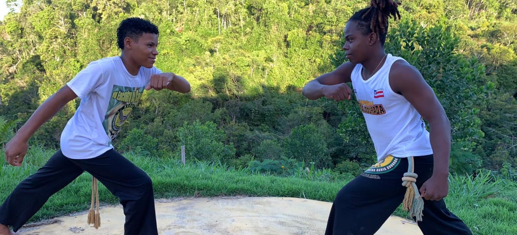 Daniel (l.) und Caíque beim Capoeira in Brasilien 2 Jungen im weißen Shirt und schwarzer Hose neigen sich mit angewinkeltem Arm einander zu. Im Hintergrun Regenwald