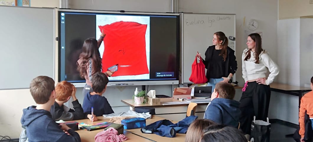 Drei weibliche Jugendliche stehen vor einer Klasse. Sie zeigen auf ein Schnittmuster an der Tafel.