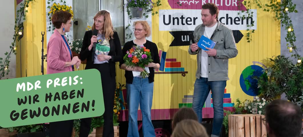 Kathrin Grau, Gründerin von Abenteuer Regenwald, nimmt den Preis entgegen, Sie steht zwischen der Laudatorin und einem Moderator vor einem mit Blumen dekorierten Bauwagen.