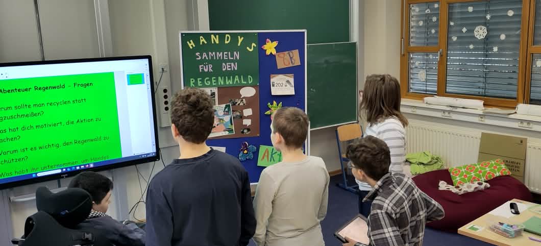 Schülerinnen und Schüler schauen auf ein Plakat mit den Worten Handys sammeln für den Regenwald.