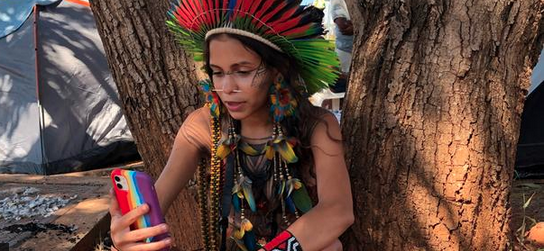 Die Aktivistin und Influencerin Alice Pataxó aus Brasilien sitzt vor einem Baum und blickt auf ihr Handy